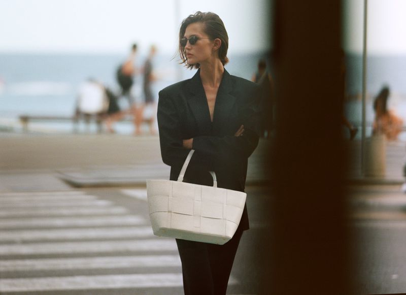 Model wearing dark sunglasses and a black blazer holding a white woven tote bag outdoors near a coastal street.