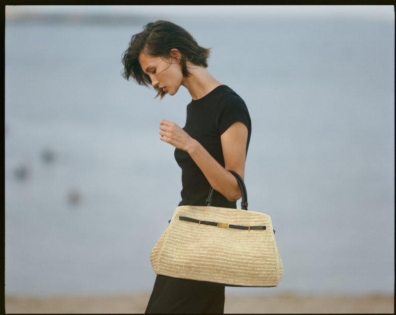 Woman in a black top holding a straw tote bag against an outdoor water backdrop