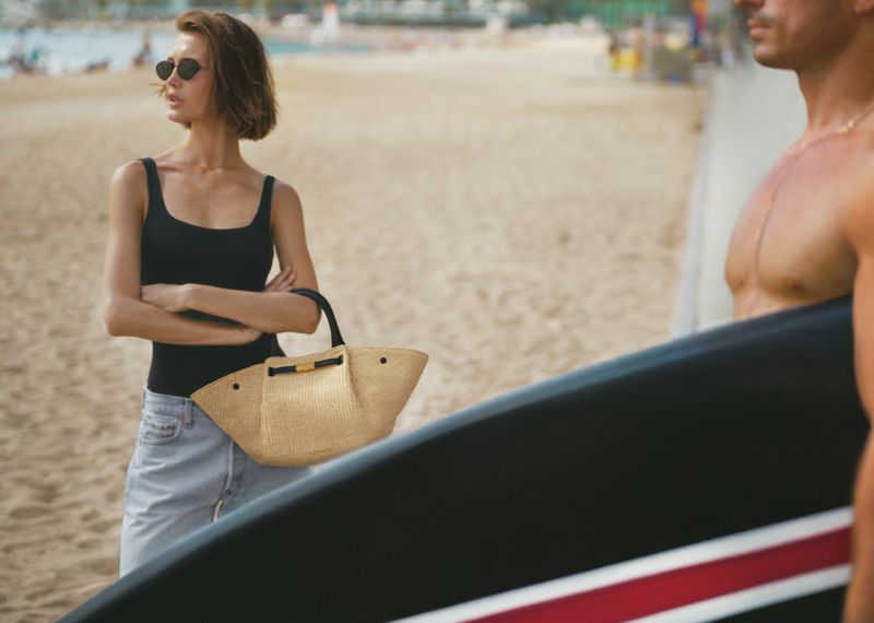 Woman in black top and sunglasses holding a straw handbag on a sandy beach next to a man with a surfboard.