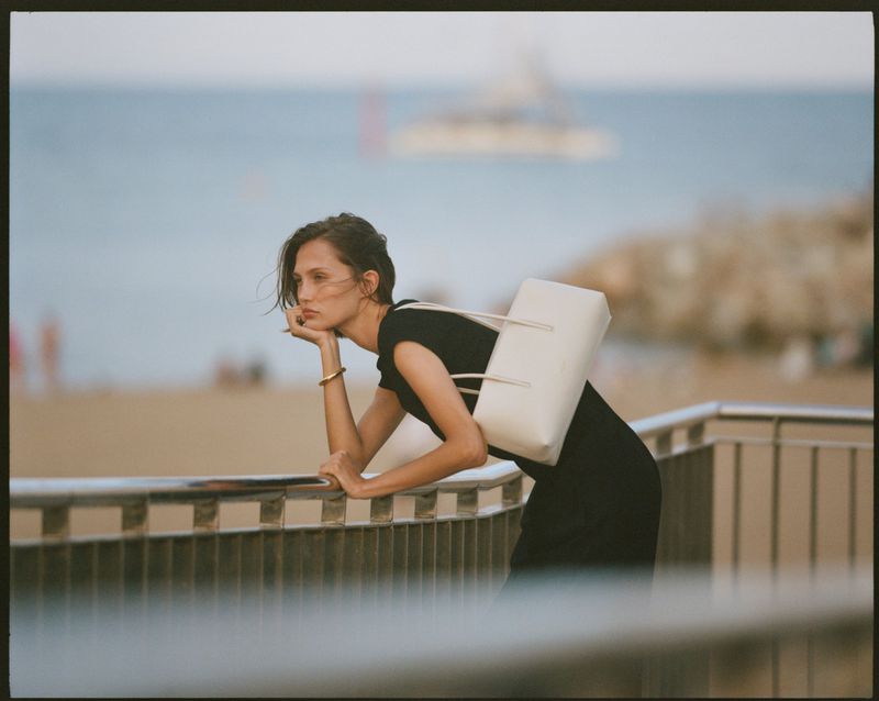 Model leaning on a railing by the ocean holding a white shoulder bag for DeMellier London