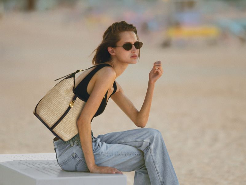A brunette woman sits on a white surface wearing sunglasses, jeans, and a black top, with a straw tote bag slung over her shoulder.