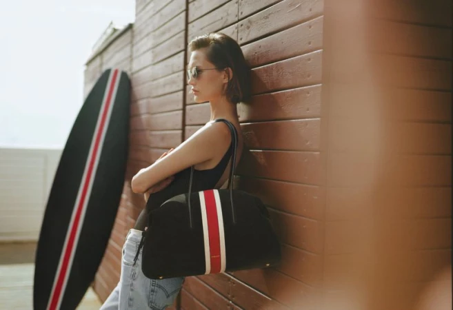 Model leaning against a wooden wall holding a dark tote bag next to a surfboard for DeMellier London