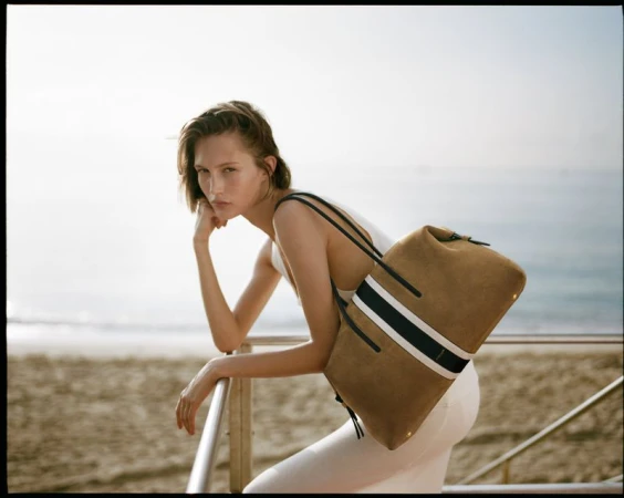 Model leaning on a railing at the beach wearing a white top and carrying a tan suede tote bag