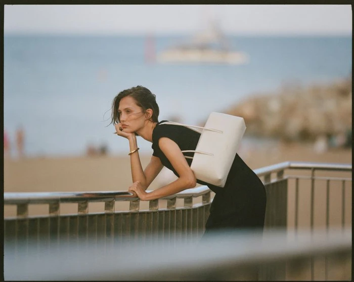 Model leaning on a railing by the ocean holding a white shoulder bag for DeMellier London