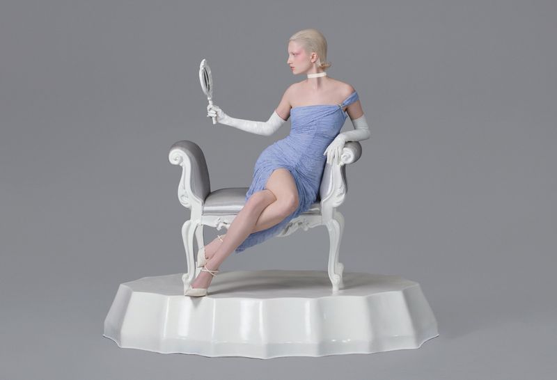 Blonde woman in blue dress seated on chair looks into hand mirror in a grey photo studio with soft lighting and visible textures.
