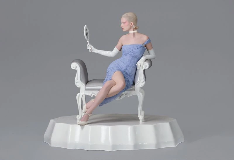Blonde woman in blue dress seated on chair looks into hand mirror in a grey photo studio with soft lighting and visible textures.