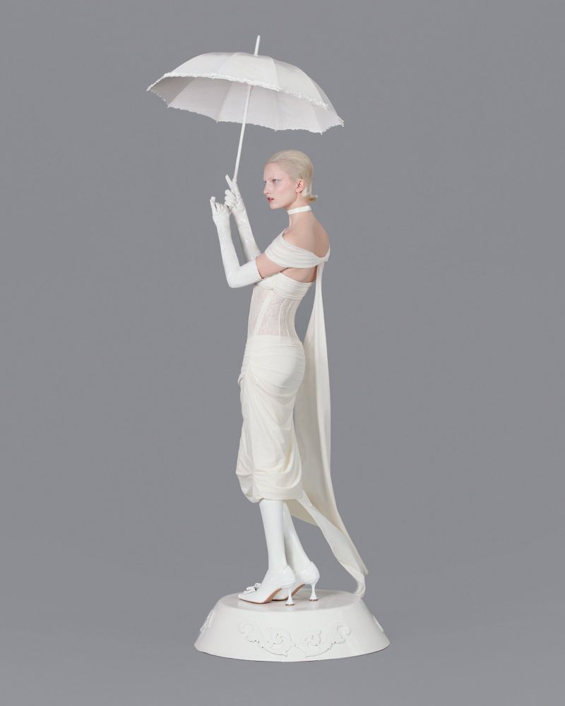 Elegant woman in white dress, wearing gloves, stands with matching umbrella on a gray backdrop. Fashion editorial photography.