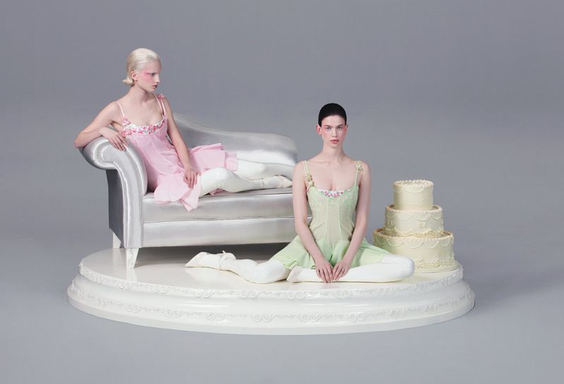Two women in light dresses pose on a platform with a cake and a chaise lounge in a studio setting, illuminated by soft, diffused light.