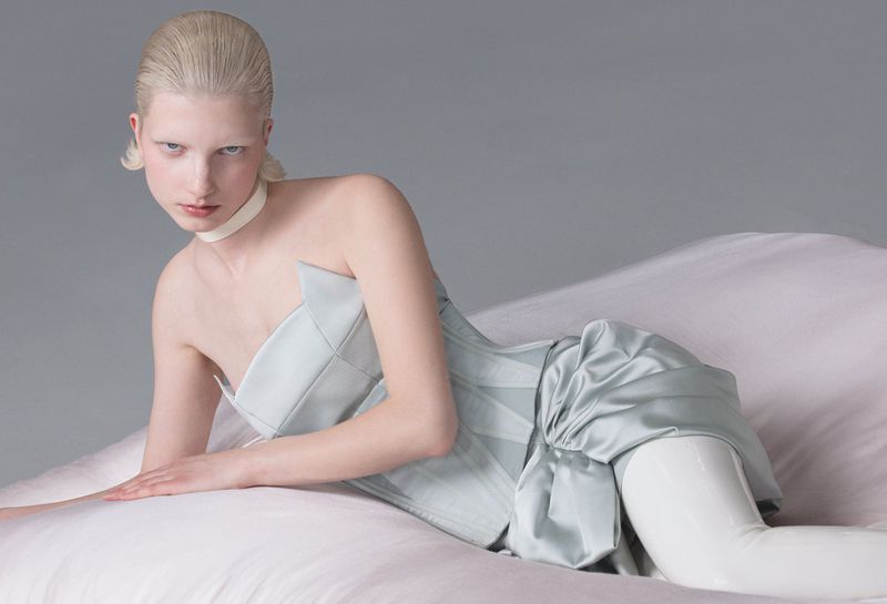 Young woman with platinum hair lies on a white surface wearing a light-blue corset, a white collar, and white leggings, in a studio setting.