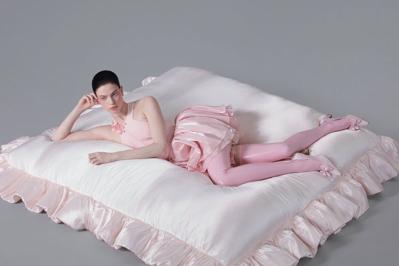 Woman in pink dress and pink stockings lying on a large white cushion, posed in a studio setting with neutral gray background.