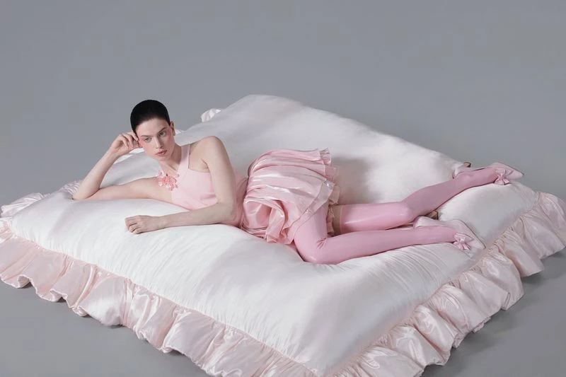 Woman in pink dress and pink stockings lying on a large white cushion, posed in a studio setting with neutral gray background.