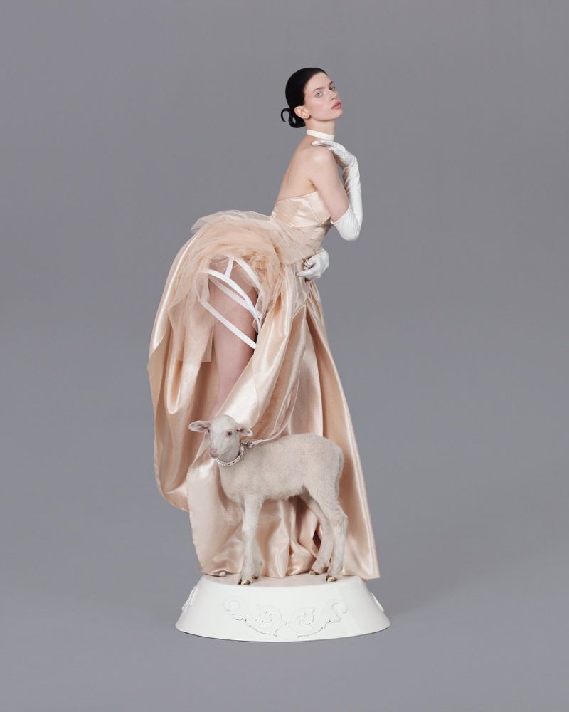 An adult female in a champagne-colored gown poses with a small lamb in a studio setting with a neutral gray backdrop.