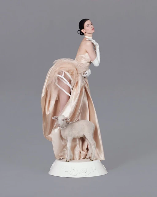 An adult female in a champagne-colored gown poses with a small lamb in a studio setting with a neutral gray backdrop.