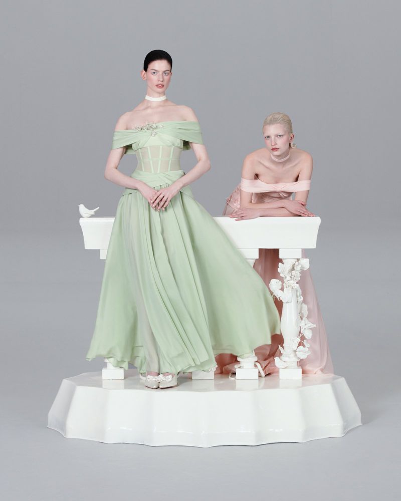 Two women in elegant off-the-shoulder dresses stand on a white platform next to a white decorative ledge in a neutral grey studio setting.