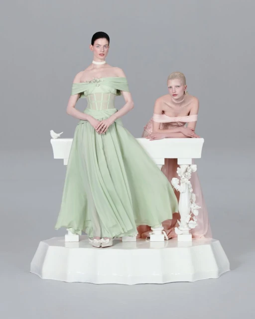 Two women in elegant off-the-shoulder dresses stand on a white platform next to a white decorative ledge in a neutral grey studio setting.