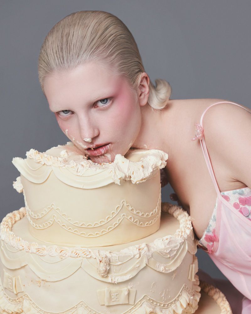 Woman with fair complexion poses while covered in cake frosting, showcasing her face, shoulders, and the two-tier frosted cake.