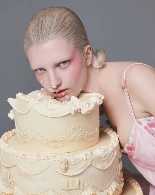 Woman with fair complexion poses while covered in cake frosting, showcasing her face, shoulders, and the two-tier frosted cake.