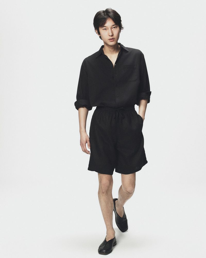 Asian man walking forward in a black shirt, black shorts, and black shoes against a plain white seamless background.
