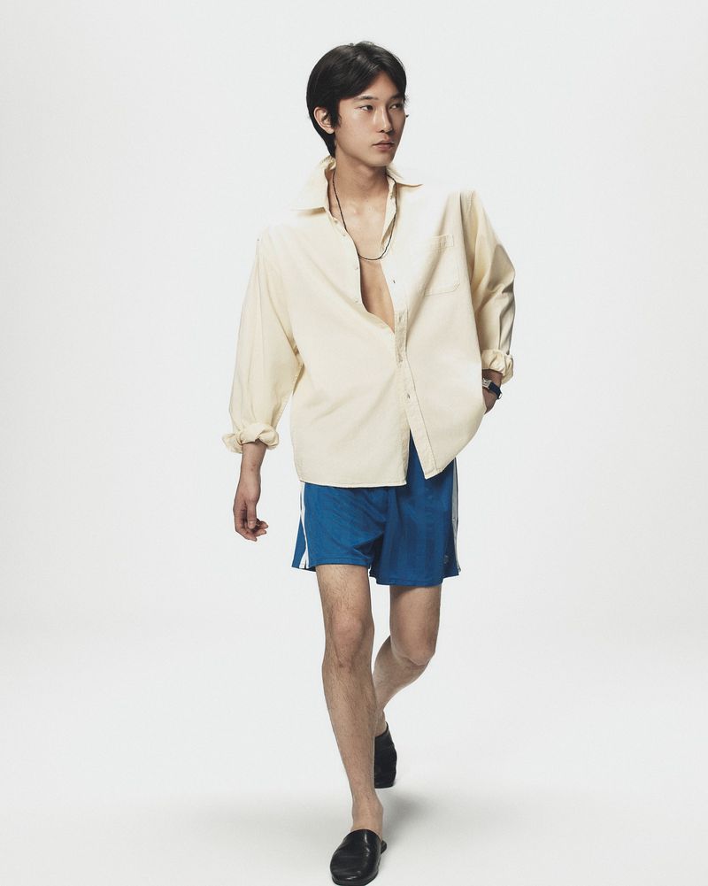 Asian man walking in cream button-up shirt and blue shorts against white background