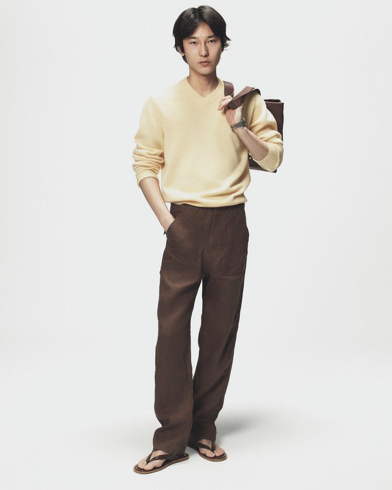 Full-length portrait of a male model wearing a pale yellow sweater, brown trousers, and dark sandals against a clean white background.