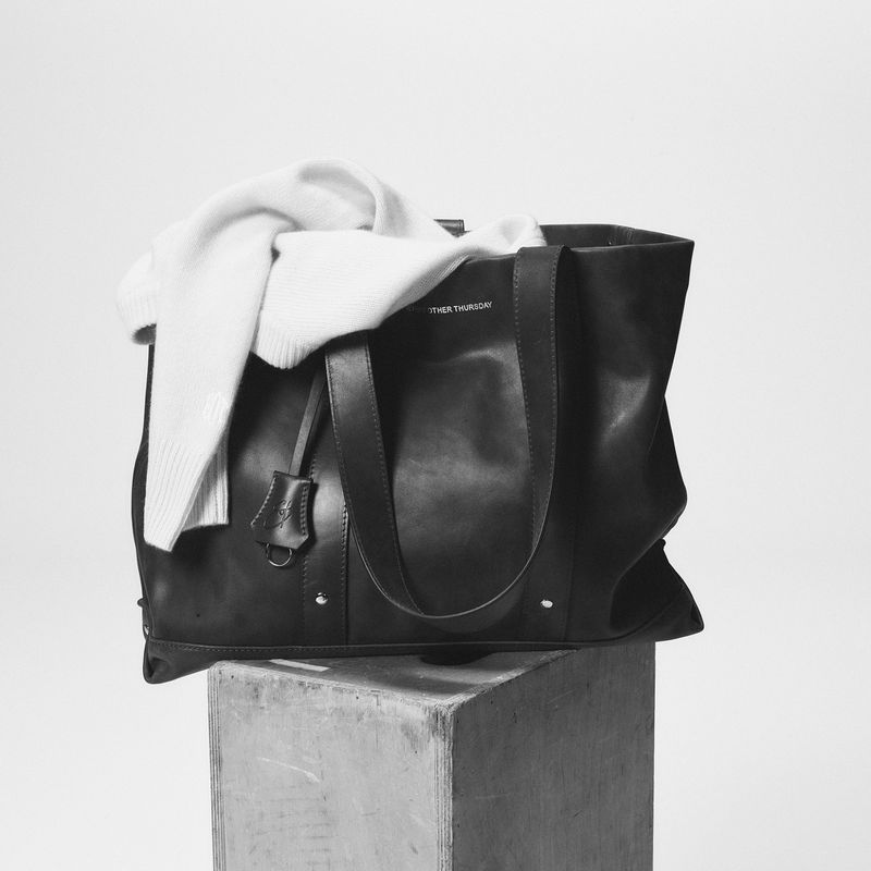 Black leather tote bag resting on a block with a white sweater draped over it against a light background.
