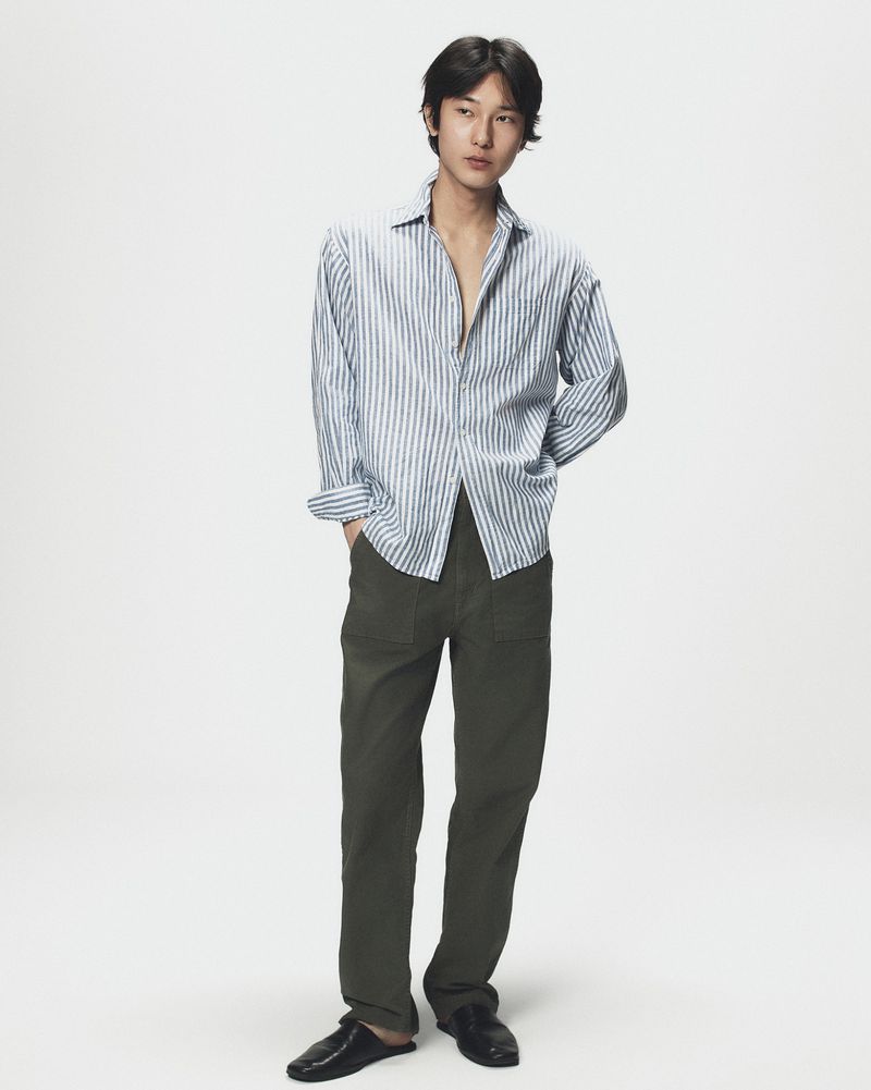 Full-length portrait of a young man wearing a blue and white striped shirt and dark green trousers against a plain light background