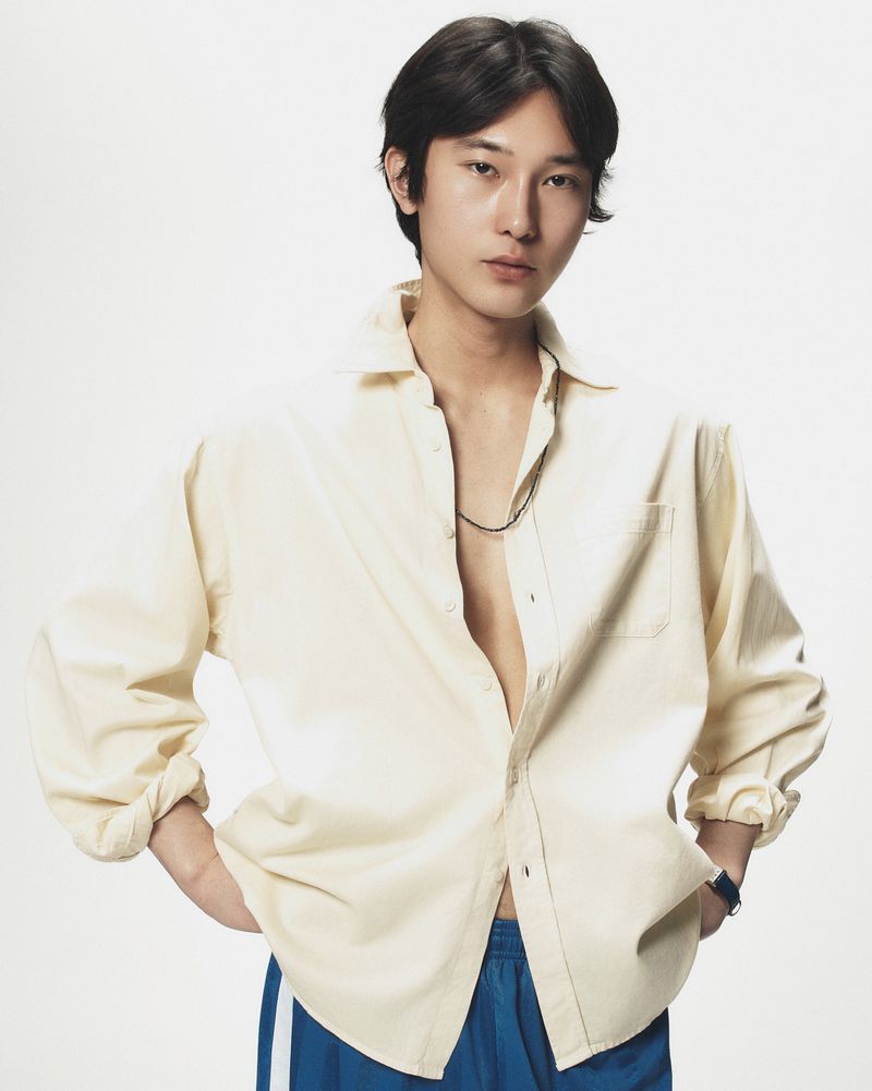 East Asian male model wearing a cream button-down shirt and blue shorts against a seamless white studio background.
