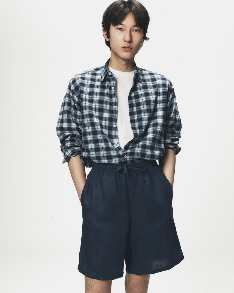 Young adult in an unbuttoned plaid shirt and dark shorts stands in a brightly lit photo studio with his hands in his pockets.
