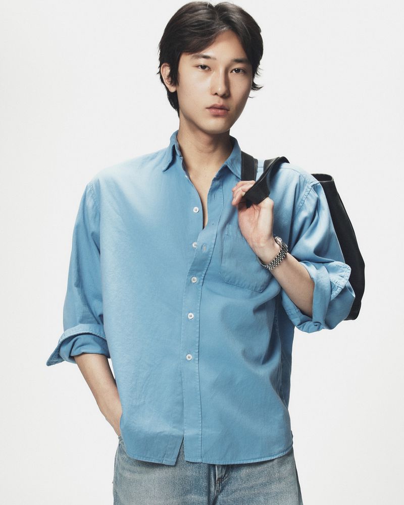 East Asian man wearing a blue button-down shirt and carrying a black bag against a white background