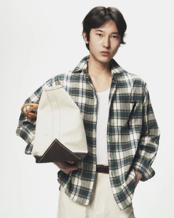 Young man in a green plaid shirt holding a canvas tote bag against a neutral background