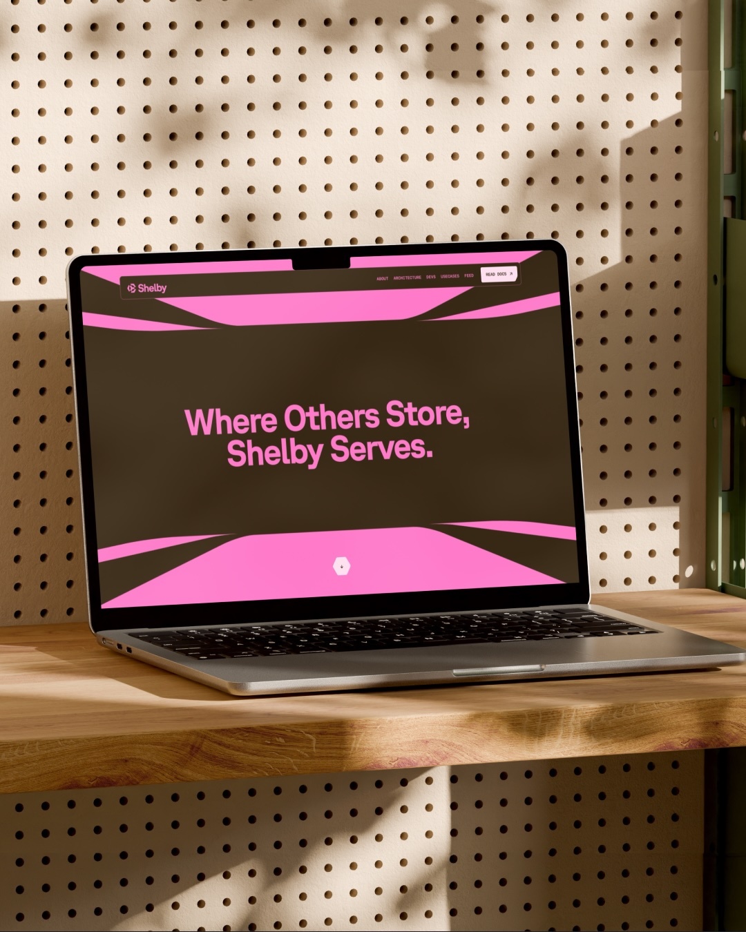 A laptop computer displaying the Shelby brand website resting on a wooden shelf against a perforated wall.