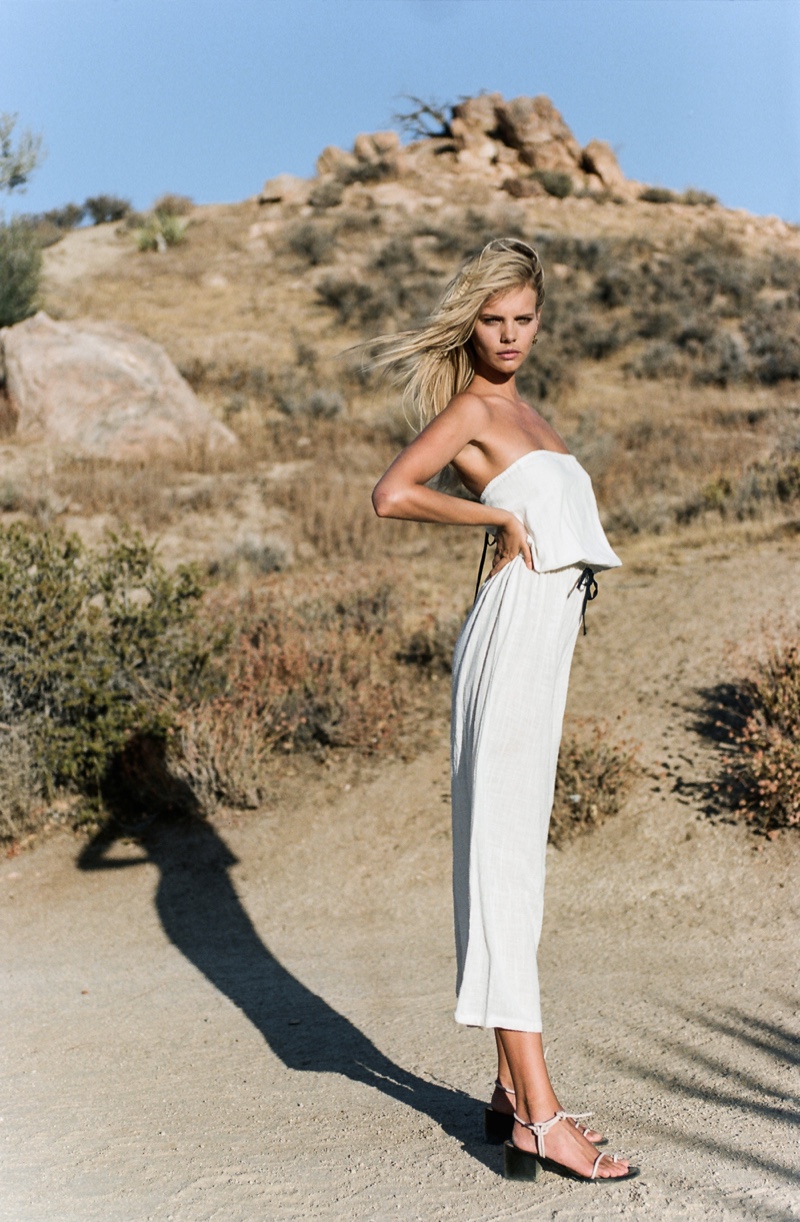 An adult blonde woman is standing in a desert wearing an off-white strapless top and pants with a mountainous landscape in the background.