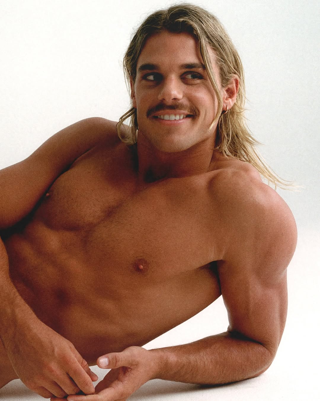 Portrait of an adult male with long hair and a mustache posing shirtless against a white background, showcasing his tanned skin.