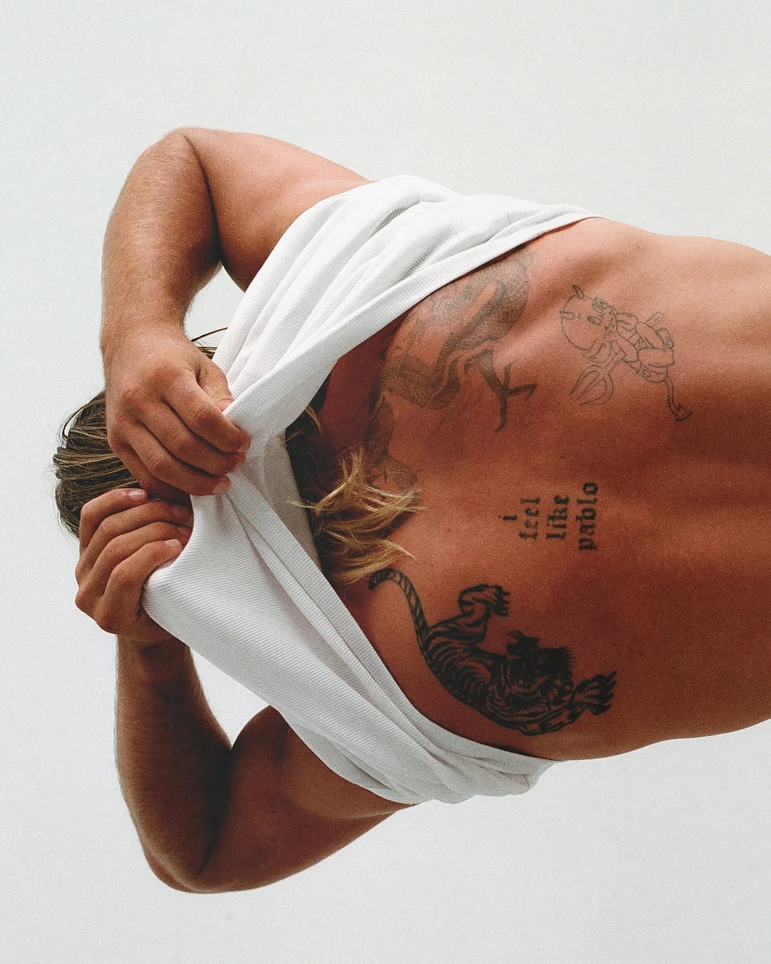 Tattooed man takes off white t-shirt, revealing upper back and blonde hair. Clean, minimalist studio shot of Caucasian adult male.