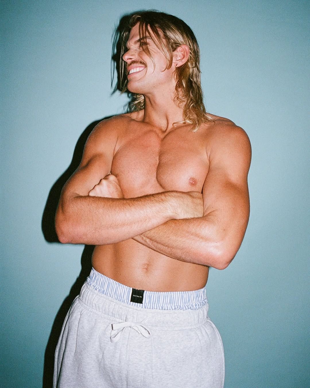 Shirtless man with long blonde hair and a mustache standing with his arms crossed in front of a blue wall, wearing grey sweatpants.