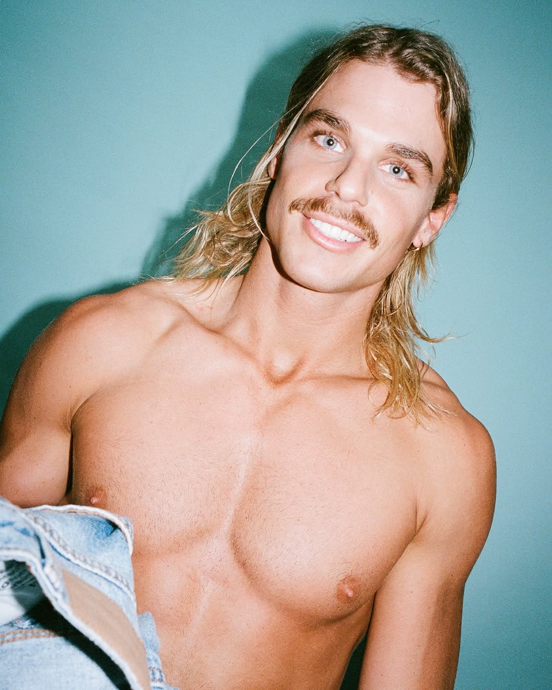 Close-up studio portrait of a fair-skinned, shirtless male model with blond hair, mustache, and blue eyes, posing against a blue backdrop.