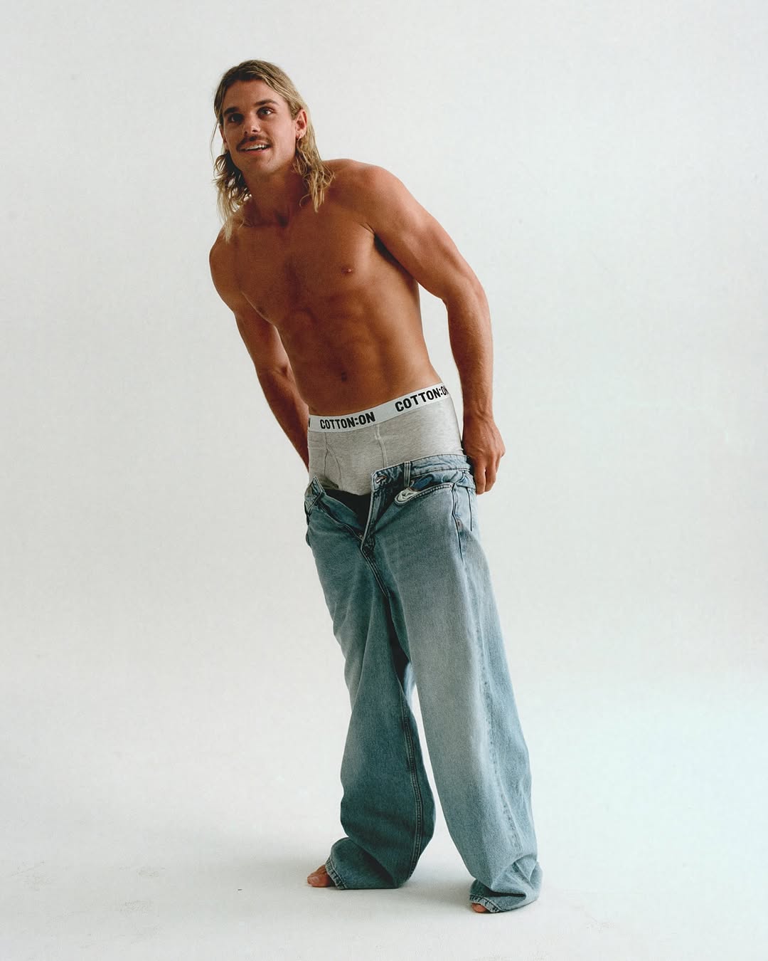 Shirtless man poses in a photo studio with jeans around his knees, revealing branded underwear, as he smiles in a full shot.