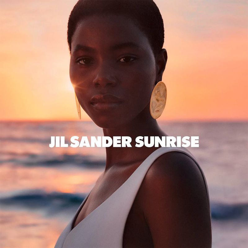 A dark-skinned woman with short hair and gold disc earrings poses in front of an ocean sunset. She wears a white top. Fashion shot.