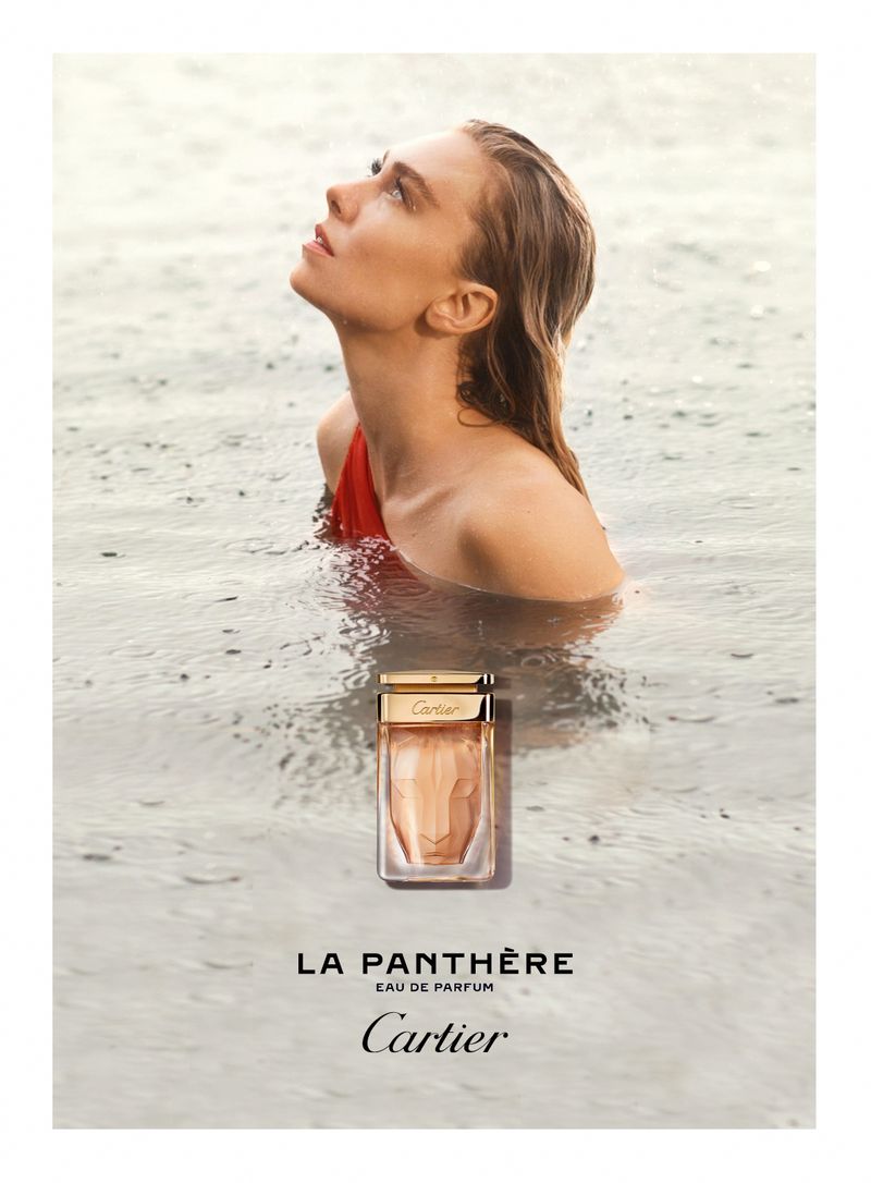A blonde woman wearing a red top stands waist-deep in water near a bottle of La Panthère Cartier Eau de Parfum in an outdoor setting.