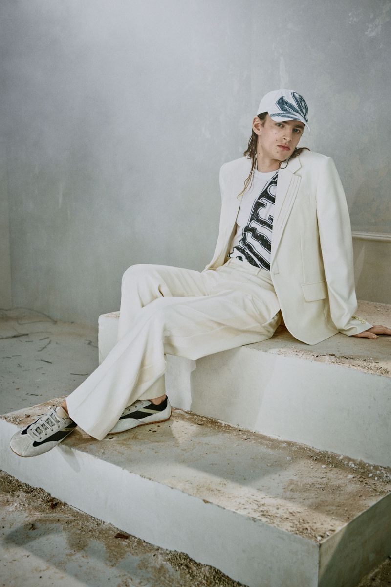 Young adult in blazer, t-shirt and baseball cap sits on a concrete structure. Fashion shot in muted indoor environment.
