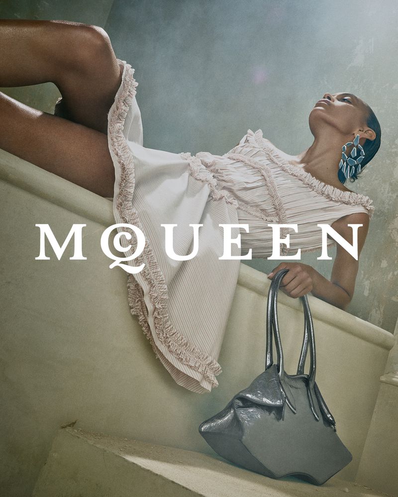 Model reclining in a ruffled dress holding a metallic handbag against an architectural background for Alexander McQueen.