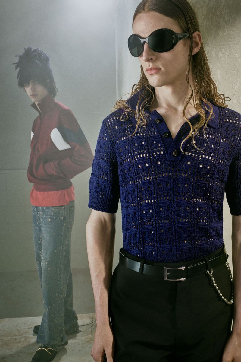 Model in blue crochet polo shirt and dark sunglasses with a second model standing in the background.