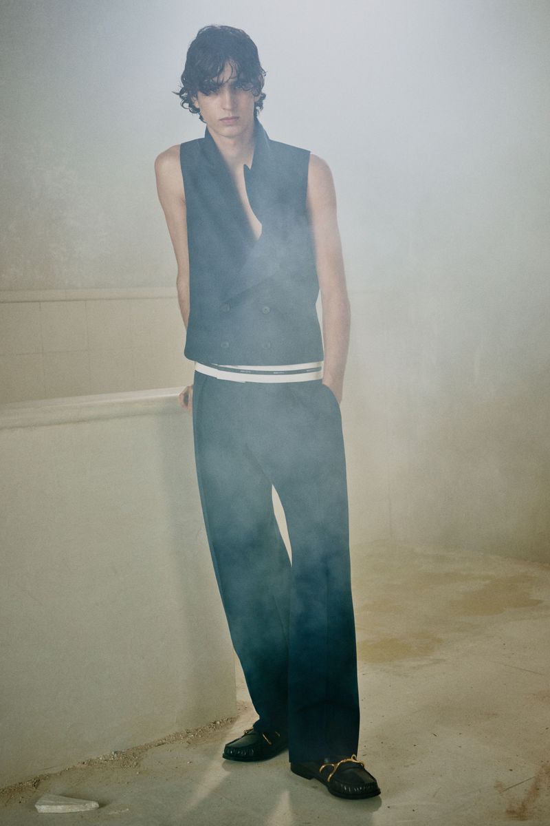 Model standing in a sleeveless vest and wide trousers against a hazy, neutral textured background.