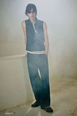 Model standing in a sleeveless vest and wide trousers against a hazy, neutral textured background.