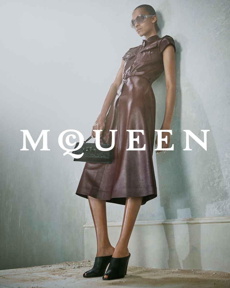 Model in dark sunglasses and brown belted dress leaning against a pale wall holding a black handbag
