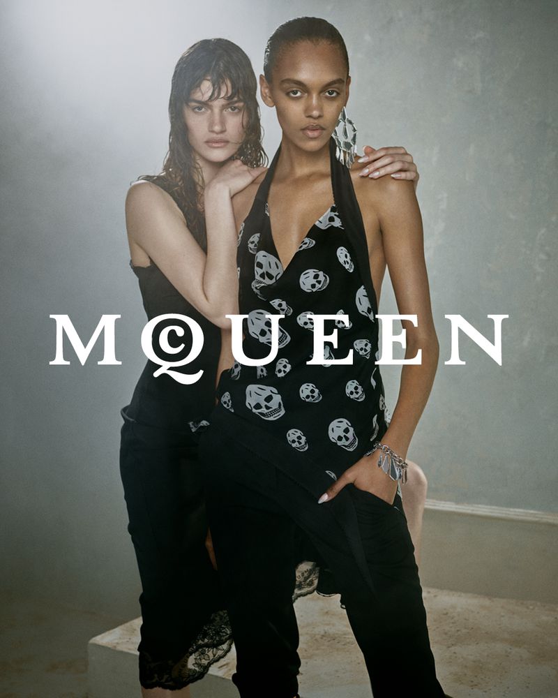 Two female models posing against a textured background wearing black clothing with graphic skull patterns for Alexander McQueen