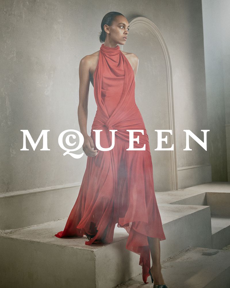Model in a draped coral red halterneck dress standing on concrete steps against a neutral architectural archway wall
