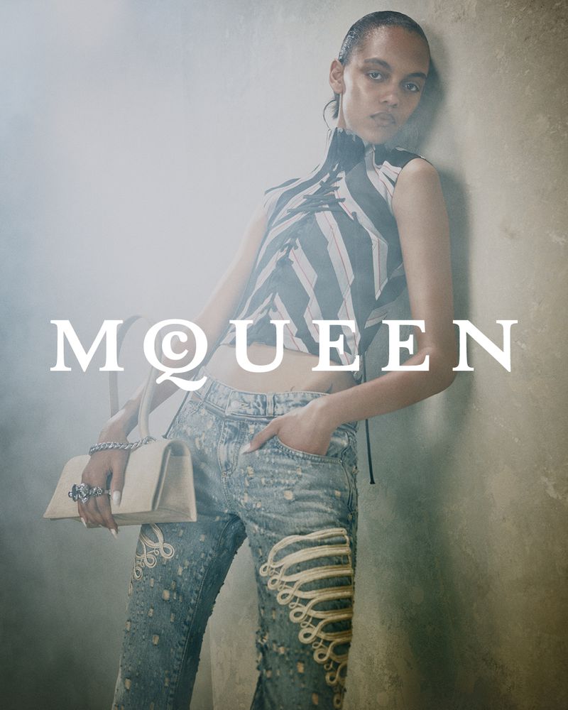 Model leaning against a textured concrete wall wearing a patterned top and distressed denim jeans holding a structured handbag.