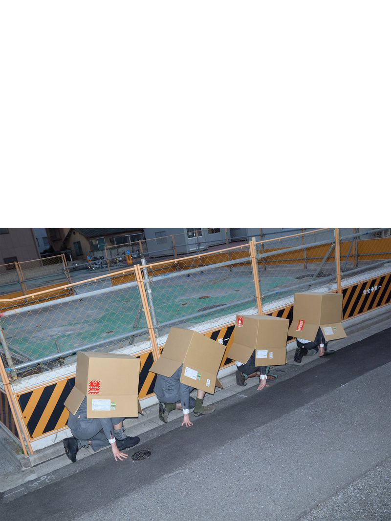 Four people in a line crouch outdoors wearing cardboard boxes over their heads against a barrier and road.