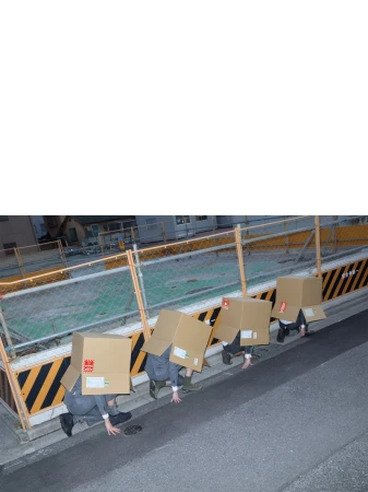 Four people in a line crouch outdoors wearing cardboard boxes over their heads against a barrier and road.